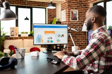 Uzaktan kumandalı erkek yönetici ev masasında oturmuş, mali verileri düzenliyor ve şirket ilerleme raporlarını bilgisayarda analiz ediyor. Afrikalı Amerikalı adam küçük işletmeler için finansal stratejiler formüle ediyor.