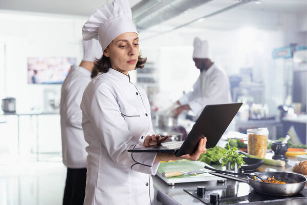 Young chef in restaurant kitchen combines traditional cookery with digital tools, using laptop to discover new recipe dish while preparing fresh produce. Female expert cooking on the stove.