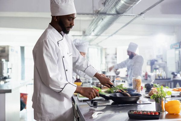 African american chef cooking wagyu beef steak with tongs in a pan, working on a delicious juicy meal at the restaurant kitchen counter and stove. Young man preparing fried food, chef life.