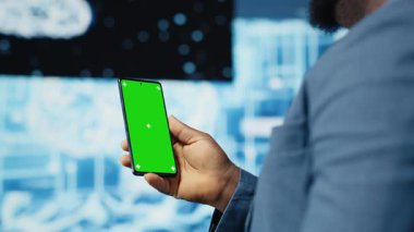 Odaklanmış siyah bilişim uzmanı, dijital yapay zeka ortamında duran yeşil ekranlı telefona bakıyor. Büyük veri otomasyonu için derin öğrenme görevleri çözmek. Sibernetik. Kamera B.