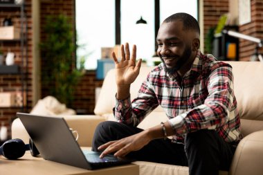 Kanepede oturan, cihaz ekranına el sallayan, online iş kursuna katılan mutlu serbest meslek sahibi bir adam. Uzaktan kumandalı siyah erkek yönetici oturma odasından iş arkadaşlarıyla neşeyle etkileşime geçiyor..