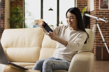 Kendine güvenen serbest çalışan bir kadın akıllı telefonda video oyunu oynarken koltukta dinleniyor. Mobil cihazlı Asyalı bayan girişimci, rahat tuğladan duvarlı oturma odasında işe ara verip eğleniyor..