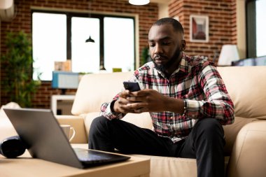 Afro-Amerikan iş adamı evden çoklu görev yapıyor, akıllı telefon kullanmak için dizüstü bilgisayarını durduruyor. Siyah erkek girişimci kişisel ve iş güncellemelerini internet üzerinden yönetirken bağlı kalıyor.
