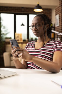 Gözlüklü siyahi bir kadın, dizüstü bilgisayarla masada oturuyor, telefon bildirimlerini ve sosyal medyayı kontrol ediyor. Afrika kökenli Amerikalı uzaktan kumandalı yönetici cep telefonuyla iş arkadaşlarıyla sohbet ediyor.