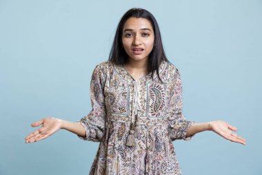 Genç bir yetişkin, kameranın önünde bir jest bilmediğimi söylüyor. Bir soruya cevap vermek için cahil ve cahil gibi davranıyorum. Unutkan güvensiz kadın mavi arka planda şaşkın hissediyor.