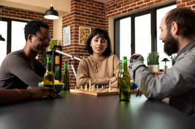 Kollarını kavuşturmuş beyaz bir kadın, erkek arkadaşlarının arasında oturmuş tuğla duvar odasında satranç oynuyor. Diğerlerinin oyun oturumundan zevk alırken kameraya bakan genç bayana odaklan.