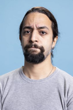 Stüdyoda kollarını kavuşturarak poz veren hayal kırıklığına uğramış genç bir adam mavi arka planda ciddi bir şekilde giyinmiş. Uzun kıvırcık saçlı ve kamerada sakalı olan genç biri..