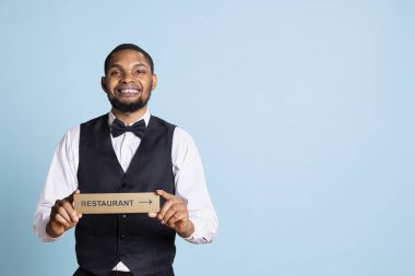 Kapıcı otel tabelasıyla restoranın yönünü gösteriyor. Konuklara konforu gösteren bir gösterge tutuyor. Duvar izi kullanarak takım elbiseli ve kravatlı profesyonel otel kapıcısı..