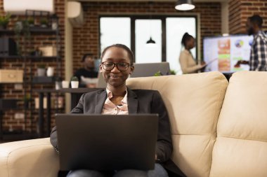 Gülümseyen siyah kadın girişimci rahat ofis salonundan çalışır, bilgisayarla iş verilerini analiz eder. Profesyonel kadın e-postalarını kontrol ediyor ve kişisel bilgisayarıyla verimli bir güne hazırlanıyor..