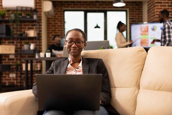 Smiling black female entrepreneur works from cozy office lounge, using laptop to analyze business data. Professional woman checking emails and preparing for productive day with her personal computer.