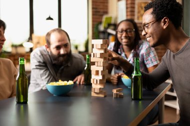 Gözlüklü genç siyahi adam, kapalı oturum sırasında Jenga Kulesi 'nden ahşap bloğu mutlulukla çekiyor. Çeşitli arkadaş grupları dikkatle izliyor, tuğla duvar odasında paylaşılan anların tadını çıkarıyorlar..