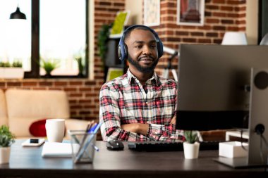 Siyah erkek serbest çalışan, kulaklıkları ve kolları bağlı bir şekilde masaüstü bilgisayarında internetten Webinar izliyor. Kablosuz kulaklık takan Afrikalı Amerikalı iş adamı bilgisayardaki video sunumunu inceliyor..