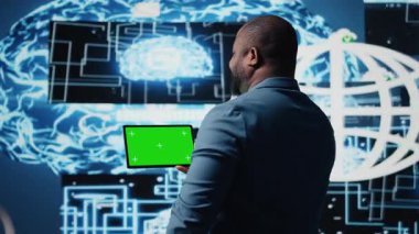 Siyahi bilişim uzmanı kroma anahtarının yanındaki algoritmik verileri inceliyor, ileri teknolojili dijital görsellerle dolu bir ortamda tablet kullanıyor. AI akıllı teknoloji ve sinirsel ağlar. Kamera A.