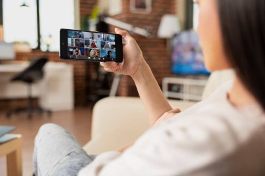 Kanepede oturan ve sanal toplantıya cep telefonuyla katılan kadın girişimcilere yakın çekim. İş kadını, çevrimiçi olarak farklı konumlardan çeşitli meslektaşlarıyla işbirliği yaparak mobil aygıtı kavrıyor.