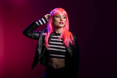 Pembe kaküllü, saç kesimi ve göz kalemi olan genç, modaya uygun bir bayan stüdyo resminde kendine güvenen bir kıyafetle ayakta duruyor. Deri ceketli ve kırbaç bluzlu, kollarını kaldırmış poz veren çekici beyaz kadın..