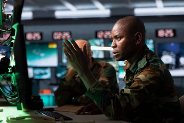 Gözetleme monitörüne bakan ordu subayı, görev ihtiyaçlarına göre kaynakları birimlere ayırmak için verileri analiz ediyor. Ekranda stratejik verileri inceleyen Afrikalı Amerikalı ajan