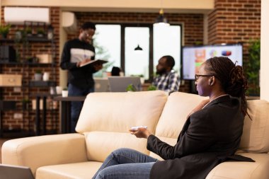 Tuğla duvardaki koltukta oturan profesyonel siyahi bir kadının projeksiyon cihazını tuttuğunu gösteren fütüristik iş etkileşimi. Modern çalışma alanı takım çalışması ve yeniliği yansıtır..