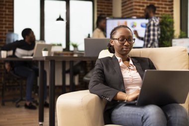 Siyah kadın şirket danışmanı, dizüstü bilgisayar kullanıyor, kanepeye oturmuş, pazarlama stratejisini analiz ediyor ve raporları derliyor. Afrika kökenli Amerikalı kadın yönetici, yeni açılan ofiste rahatça iş projesinde çalışıyor.