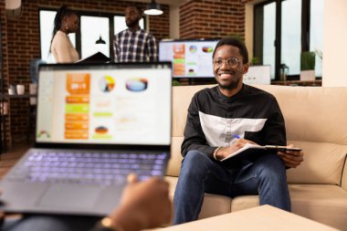 İki iş adamı, fikirlerini paylaşmak ve proje görevinde işbirliği yapmak için ofiste bir araya geldiler. Afro-Amerikan satış müdürü dizüstü bilgisayarla koltukta oturan şirket müşterisine ticari teklif sunuyor..