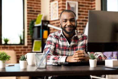 Masasında tuğla duvarlı oturma odasında bilgisayarı olan serbest çalışan siyahi bir adamın portresi. Kendinden emin Afrikalı Amerikalı girişimci kameraya gülümsüyor, uzaktan çalışma sorumluluklarına hazır..