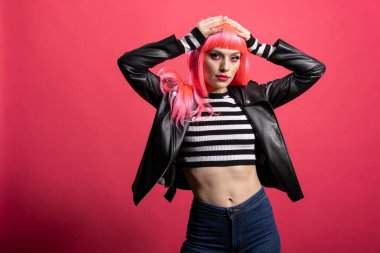 Pembe saçlı ve kalın ceketli modacı, izole edilmiş arka planda kendine güvenen rockçı tarzını benimsiyor. Güzel bir kadın hoş bir tutum sergiliyor ve zarif bir şekilde duruyor. Punk havasını ve şık havasını yansıtıyor..