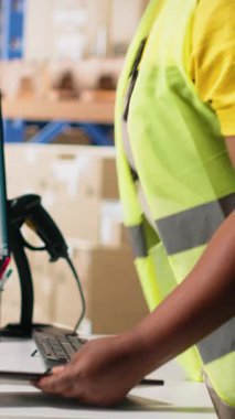 Dikey Afrikalı Amerikalı işçiler depo ortamında e-ticaret kargo siparişlerini işliyor, çelik yelek giyiyor ve paketsiz dağıtımı garantiliyor. Büyük raflarda depolanmış karton kutular