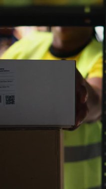 Dikey Video Tamamlama Merkezi personeli sipariş takibi için havayolu faturalarını tarar, depodaki nakliye etiketlerinde tarayıcı kullanır. Depoda raflı yelek giyen bir işçi.