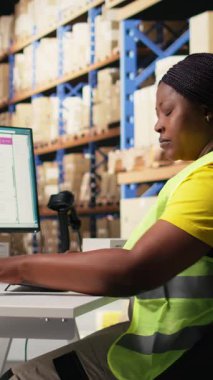 Dikey Video Afrikalı Amerikalı çalışan sipariş lojistik yazılımını güncelliyor, sevkiyat etiketlerini dağıtım için hazırlıyor. Ticaret lojistik tesisinde çelik yeleği olan endüstriyel depo personeli.