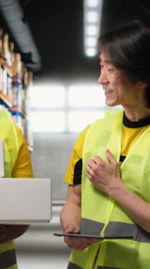 Dikey Afrikalı Amerikalı ve Asyalı ekip kargo mallarını canlı yayında sunuyor ve rafların yakınındaki endüstriyel depoda bir video kaydediyor. Büyük iş reklamları için içerik. Kamera.