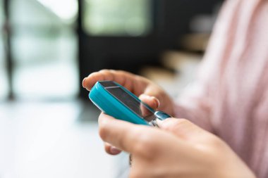 Hemşire kan şekerini glukometre cihazıyla ölçüyor. Hastane bekleme odasında rutin sağlık kontrolü yapıyor. Asyalı kadın destek alıyor ve glikoz seviyelerini takip etmeyi öğreniyor. Kapat..