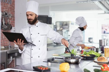 Mutfak uzmanı dizüstü bilgisayardaki yeni tarifleri araştırıyor ve ticari mutfakta çalışarak taze ürünlerle gurme yemekleri hazırlıyor. Yenilikçi ve lezzetli mutfak, gastronomi.
