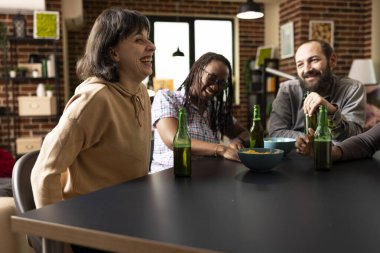 Kahkahalar ve şakalarla kaynaşan, rahat bir yaşam alanında atıştırmalık ve içeceklerle çevrili çok ırklı bir arkadaş grubu. Kafkasyalı kadın yakınlarda içkiler ve atıştırmalıklarla komik bir hikaye paylaşıyor..