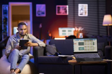 Genç Siyahi Adam kanepede rahatlıyor ve PC ile hisse senedi alım satımlarını takip ediyor, borsa grafiklerini finansal büyüme için analiz ediyor. Uzak çalışan gelir eğilimlerini ve çevrimiçi yatırımlar için ticaret verilerini gözden geçirdi.