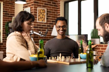 Kafkasyalı arkadaşlarıyla masa oyunu oturumunda oturan, gözlüklü neşeli siyahi bir adam. Afro-Amerikalı adam rahat tuğla duvar odasında sıradan bir satranç maçında kameraya neşeyle gülümsüyor..