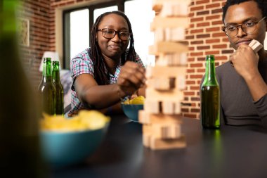 Afrika kökenli Amerikalı kadın ve erkek yakınlarda içki ve atıştırmalıklarla Jenga oyunu oynuyorlar. Endişeli kadın şahıs dikkatli bir şekilde tahta kuleden blok kaldırarak oyun sırasında dengesini koruyor.