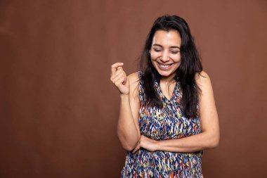 Güzel Hintli kadın kontrolsüz bir şekilde gülüyor, kahverengi arka planda tek başına duruyor. Kollarını kavuşturup poz veren neşeli kadın modern stüdyoda gülümsüyor..