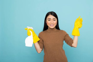Lastik eldivenli neşeli Asyalı kadın bir elinde temizlik spreyi, enerji ve pozitiflikle kollarını kaldırıyor. Gülümseyen hizmetçi mavi arka planda dezenfektan şişesi tutuyor.