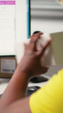 Dikey Video Siyah Kadın malları paketliyor ve kutulara nakliye etiketleri yapıştırıyor, kolay dağıtım için etiketlerde yanlış takip numaraları yaratıyor. Yapışkan hava yolu etiketleri kullanan bir işçi. Kamera A.