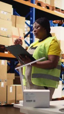 Dikey Video Siyah kadın işçi envanter kontrolü için dosyalardaki yanlış etiketleri doğruluyor, raflardaki paketleri sayıyor ve depoya hızlı teslimat sağlıyor. E-ticaret havayolu faturaları. Kamera A.