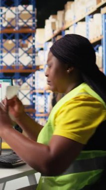 Dikey Video Siyah depo çalışanı etiketleme ve paketleme işlerini yönetir, paketlere basılı havayolu faturası takip numaraları etiketleri uygular. Yelekli kadın depoda işlem emri veriyor.