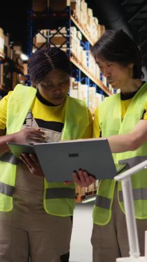 Sanayi deposundan gelen raflı kargo sevkiyatını takiben çeşitli çalışanlardan oluşan dikey video ekibi tablet üzerindeki parsel durumu için izleme yazılımı kullanıyor. Çelik yelek personeli. Kamera A.