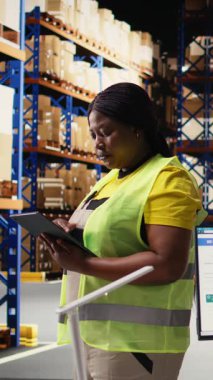 Dikey Video Siyah depo çalışanı, teslimat servisi için envanter kontrolü için tablet üzerinde paket izleme arayüzü kullanıyor, büyük depolama raflarıyla dolu endüstriyel depo. İnternetten alışveriş