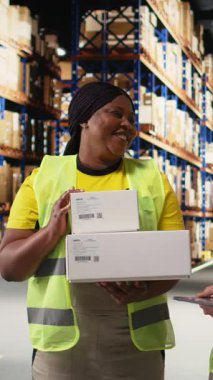 Dikey Afrikalı Amerikalı ve Asyalı ekip kargo mallarını canlı yayında sunuyor ve rafların yakınındaki endüstriyel depoda bir video kaydediyor. Büyük iş reklamları için içerik. Kamera.