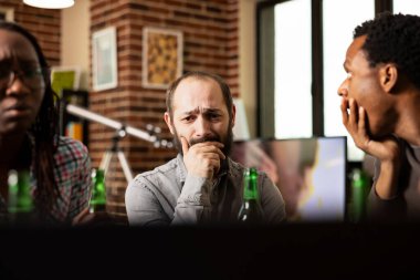 Endişeli beyaz adam, Afrika kökenli Amerikalı arkadaşlarıyla duvar odasında duygusal içeriği izliyor. Masada oturan sakallı erkek, üzgün görünüyor. Ağzı kapalı hüzünlü videolar izliyor.