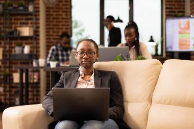 Siyahi kadın yönetici rahat bir koltukta oturur, başlangıç araştırma verilerini analiz etmek ve şirket raporlarını yönetmek için dizüstü bilgisayar kullanır. Gözlüklü odaklanmış iş kadını, e-postaları gözden geçiriyor ve iş projelerini hazırlıyor..