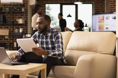 Siyah erkek çalışan bilgisayarındaki mali kayıtları güncelliyor. Başlangıç ofisindeki bir panoya atıfta bulunuyor. Odaklanmış işadamı, iş raporlarını yönetmek için dijital araçlar kullanarak şirket evraklarını gözden geçiriyor.