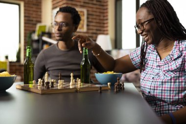 Satranç oynayan neşeli siyahi kadın, bir taşı hareket ettirirken heyecanla gülümsüyor, kazanmayı bekliyor. Gözlüklü Afrikalı Amerikalı kadın, arkadaşlarıyla içeride oyun oynuyor..