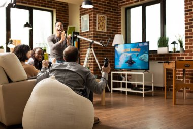 Çok ırklı arkadaşlar, oturma odasındaki kanepede toplanan modern oyun konsolunda oynanan uzay simülasyon oyununu kazandıktan sonra sevindiler. Kahkahalar ve kutlamalar, takılma sırasında duvarları ören duvarlarla doluydu..