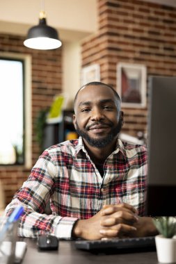 Sırıtan siyah uzaktan kumandalı bir işçi, masaüstü bilgisayarı ve tuğla duvarlı rahat bir yaşam alanı olan bir masada oturuyor. Serbest meslek sahibi erkek profesyonel evden sanal toplantılar ve iş görevleri için hazırlandı.