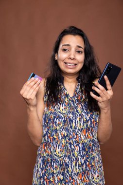 Banka kartının ifşa olduğunu anladıktan sonra elinde akıllı telefon tutan endişeli bir kadın. Gergin ve üzgün olan kadın, çevrimiçi dolandırıcılık ya da dolandırıcılığın kurbanı olmaya tepki veriyor..